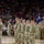 A military formation ceremony is taking place on the floor of an indoor sports arena, with soldiers in OCP (Operational Camouflage Pattern) uniforms standing at attention in organized rows. Two blue guidon flags bearing the number '158' are visible, indicating a unit designation. A large crowd of civilians fills the arena seating, suggesting this is a public send-off, homecoming, or activation ceremony.