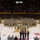 A large military ceremony is taking place inside what appears to be Arizona State University's Wells Fargo Arena (now Desert Financial Arena), identifiable by the maroon and gold court markings and PAC-12 logo. Hundreds of soldiers in camouflage uniforms are assembled in formation on the basketball court floor, with a color guard presenting flags at the center. The arena stands are filled with a large civilian audience observing the event.