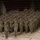 A large formation of military personnel in Army Combat Uniforms (ACU/OCP pattern) are rendering a salute in unison inside what appears to be an indoor sports arena or gymnasium. The formation is arranged in tiered rows, with a guidon bearer visible at the front center holding a blue unit flag. This appears to be a formal military ceremony such as a deployment, redeployment, or unit activation event.