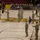 A military ceremony is taking place on the basketball court of Arizona State University's Sun Devil Arena. Soldiers in camouflage uniforms are standing in formation while holding unit guidon flags, with an audience of military personnel and civilians seated in the stands applauding. The event appears to be a formal military function such as a change of command or unit activation ceremony.