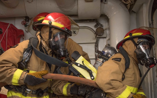 A team of firefighters in full protective gear, including tan/yellow turnout suits, red helmets, and SCBA (Self-Contained Breathing Apparatus) face masks, are maneuvering through a narrow corridor or hatchway. They appear to be conducting a firefighting drill or operation aboard a naval vessel, indicated by the metal bulkheads, pipes, and 'FIREMAIN' labeling visible in the background. The team is working in close coordination, handling a fire hose together.