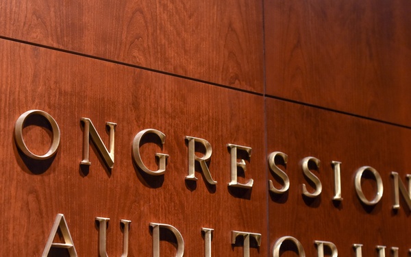The image shows a close-up of a wooden wall panel with raised metallic gold lettering reading 'CONGRESSIONAL AUDITORIUM'. The sign is mounted on rich reddish-brown wood paneling, suggesting a formal government or institutional setting. The photograph is taken at a slight angle, emphasizing the dimensional quality of the letters.