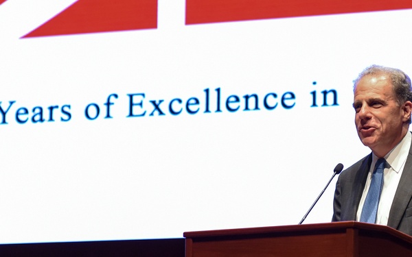 Two men are present at a formal event celebrating a 40-year anniversary. One man stands at a wooden podium speaking into a microphone, while an older man is seated nearby smiling. A large projection screen behind them displays text reading '40 Years of Excellence in [Independe...]'.