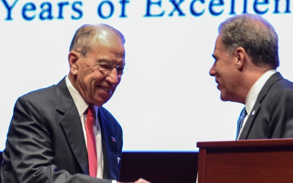 Two men in dark business suits are shaking hands on a stage near a wooden podium. The backdrop displays the text '40 Years of Excellence,' suggesting a formal anniversary or recognition ceremony. The man on the left is smiling and holding papers, while the man on the right stands near the lectern.