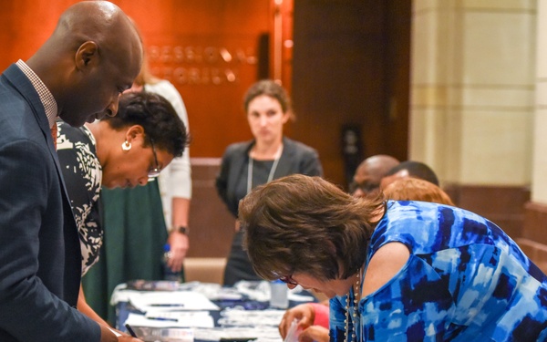 The image shows a registration or check-in table at what appears to be a professional conference or formal event. Multiple attendees are leaning over a navy blue-draped table, signing documents or collecting materials such as lanyards and name badges. The setting appears to be in a formal building lobby or hallway.