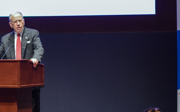 An elderly man in a dark suit with a red tie is delivering a speech at a wooden podium on a stage. Behind him is a banner for the 'Council of the Inspectors General on Integrity and Efficiency' (CIGIE), featuring red, white, and blue patriotic design elements. An audience is visible in the foreground, watching the presentation.
