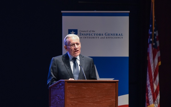 An elderly man in a dark suit is speaking at a wooden podium on a stage before an audience. Behind him is a banner for the 'Council of the Inspectors General on Integrity and Efficiency' and a United States flag. The setting appears to be a formal auditorium or conference hall.