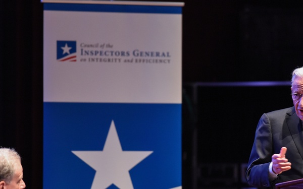 Two men are on a stage at an event hosted by the Council of the Inspectors General on Integrity and Efficiency. One man stands at a wooden podium speaking and gesturing, while another sits at a table with a microphone and water bottle. The setting appears to be a formal auditorium or conference venue.