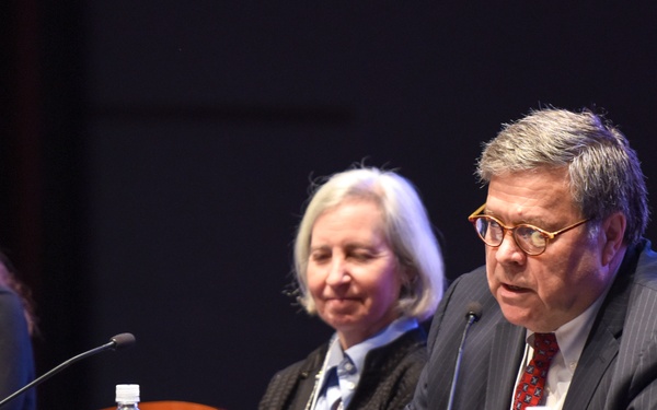 Three individuals are seated at a panel discussion table covered with a blue tablecloth. The person on the right, identified by a visible name placard as 'William Barr', is actively speaking into a microphone. The setting appears to be a formal conference or official government event.