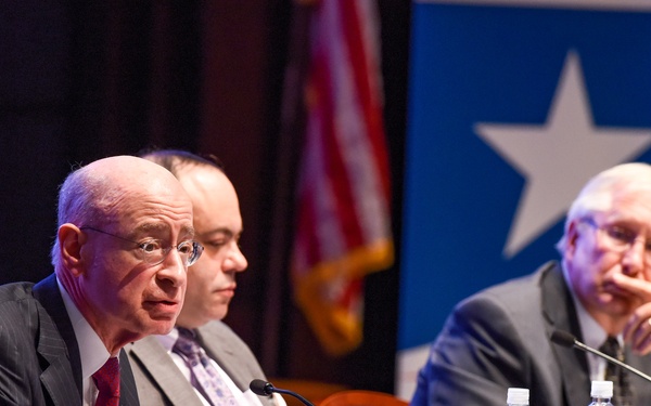 Three men in business attire are seated at a panel table covered with a blue tablecloth, appearing to be at a formal conference or hearing. The man on the left is actively speaking into a microphone with his hands gesturing, while the others listen attentively. Name placards are visible on the table, with one partially readable as 'Daniel Levinson'.