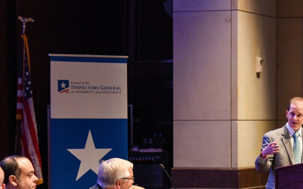 A formal panel discussion or conference hosted by the Council of the Inspectors General on Integrity and Efficiency (CIGIE). Three men are seated at a draped panel table while a fourth man in a gray suit speaks at a podium to their right. The setting appears to be an auditorium or formal meeting hall with an American flag and branded banner visible.