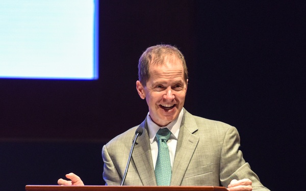 A middle-aged man in a light gray suit and teal/mint tie is speaking enthusiastically at a wooden podium on a darkened stage. He appears animated and smiling broadly, suggesting an engaging or humorous moment during his presentation. A projection screen is partially visible in the upper left background.