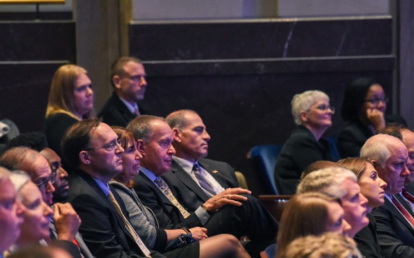 A large audience of professionally dressed individuals is seated in what appears to be a formal auditorium or conference hall, attentively watching a presentation or event. The attendees are predominantly wearing business attire including suits and formal wear, suggesting a high-level professional or governmental gathering. The venue features tiered seating with wooden chairs and marble or stone architectural elements visible in the background.