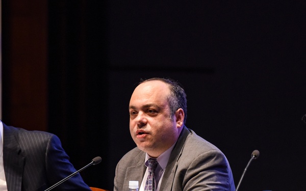 Three men in business attire are seated at a panel discussion table covered with a blue tablecloth. The central figure appears to be speaking into a microphone, with name placards visible in front of each panelist. The setting appears to be a formal conference or symposium, with institutional banners partially visible in the background.