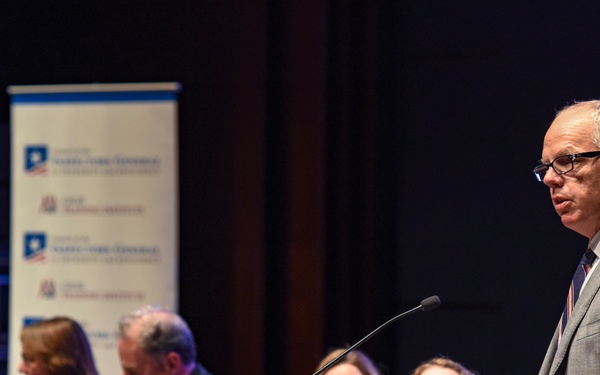A formal panel discussion or conference is taking place in an auditorium setting. A man in a gray suit stands at a wooden podium speaking into a microphone, while several panelists are seated at a draped table behind him. A branded banner is visible in the background, suggesting an institutional or academic event.