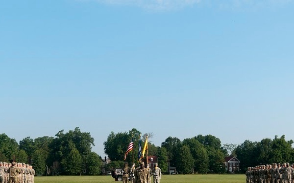 A military formation ceremony is taking place on a large open grass field, with two groups of uniformed soldiers standing in formation on either side of a central color guard bearing flags. The scene appears to be a formal military event such as a change of command, unit activation, or review ceremony. Red-brick buildings and dense green trees are visible in the background under a clear blue sky.