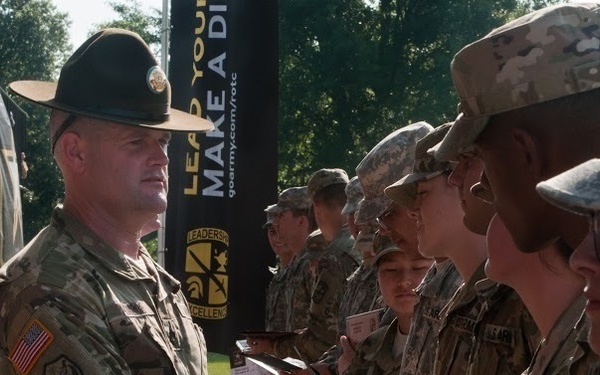 A U.S. Army officer wearing a distinctive drill sergeant campaign hat is shaking hands with soldiers in a line, appearing to present certificates or awards during a military ceremony. The event appears to be an ROTC-related recognition ceremony, as evidenced by the 'goarmy.com/rotc' banner and 'Leadership Excellence' signage in the background. Multiple soldiers in Army Combat Uniforms (ACUs) are lined up to receive recognition.
