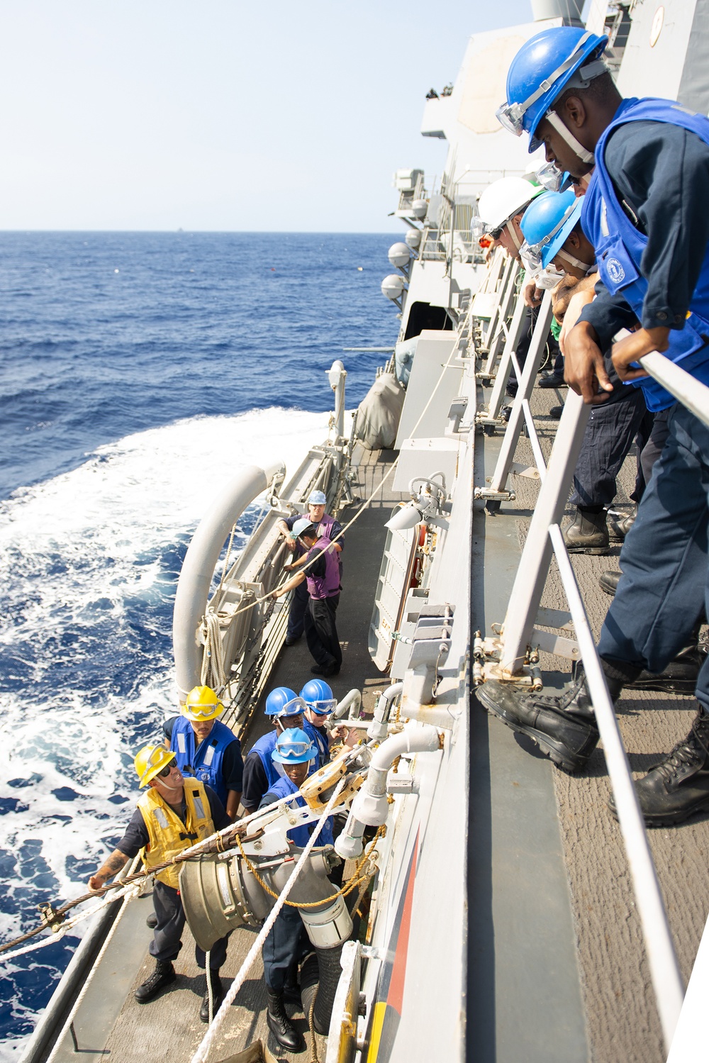 Naval personnel are conducting a replenishment-at-sea (RAS) or underway replenishment (UNREP) operation on the deck of a warship. Crew members wearing safety helmets, protective goggles, and life vests are handling lines, hoses, and rigging equipment along the ship's starboard side. The vessel is underway in open blue ocean waters under clear sunny conditions.