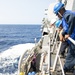 Naval personnel are conducting a replenishment-at-sea (RAS) or underway replenishment (UNREP) operation on the deck of a warship. Crew members wearing safety helmets, protective goggles, and life vests are handling lines, hoses, and rigging equipment along the ship's starboard side. The vessel is underway in open blue ocean waters under clear sunny conditions.