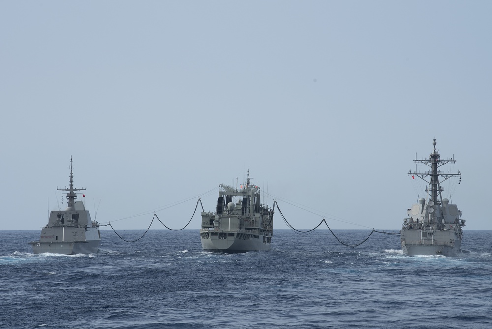 Three naval vessels are conducting an underway replenishment (UNREP) operation at sea, with a central replenishment ship simultaneously transferring supplies or fuel to two warships on either side. The ship on the left appears to be a modern frigate or destroyer with a stealth hull design, while the ship on the right displays characteristics consistent with a US Navy Arleigh Burke-class guided-missile destroyer. Replenishment lines and hoses are visibly suspended between the vessels.