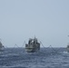 Three naval vessels are conducting an underway replenishment (UNREP) operation at sea, with a central replenishment ship simultaneously transferring supplies or fuel to two warships on either side. The ship on the left appears to be a modern frigate or destroyer with a stealth hull design, while the ship on the right displays characteristics consistent with a US Navy Arleigh Burke-class guided-missile destroyer. Replenishment lines and hoses are visibly suspended between the vessels.