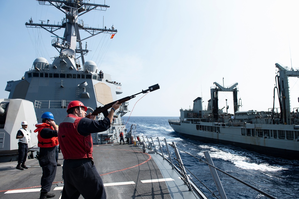 A U.S. Navy sailor wearing a red helmet and life vest is firing a line-throwing gun (shot line rifle) toward an adjacent replenishment vessel during an underway replenishment (UNREP) operation at sea. Two warships are visible — what appears to be an Arleigh Burke-class guided-missile destroyer on the left and a replenishment oiler on the right — sailing in close parallel formation. Several crew members in safety gear are observing the operation on the destroyer's deck.