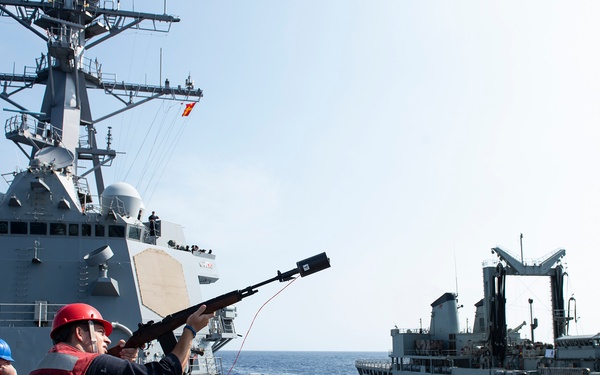 A U.S. Navy sailor wearing a red helmet and life vest is firing a line-throwing gun (shot line rifle) toward an adjacent replenishment vessel during an underway replenishment (UNREP) operation at sea. Two warships are visible — what appears to be an Arleigh Burke-class guided-missile destroyer on the left and a replenishment oiler on the right — sailing in close parallel formation. Several crew members in safety gear are observing the operation on the destroyer's deck.