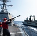 A U.S. Navy sailor wearing a red helmet and life vest is firing a line-throwing gun (shot line rifle) toward an adjacent replenishment vessel during an underway replenishment (UNREP) operation at sea. Two warships are visible — what appears to be an Arleigh Burke-class guided-missile destroyer on the left and a replenishment oiler on the right — sailing in close parallel formation. Several crew members in safety gear are observing the operation on the destroyer's deck.
