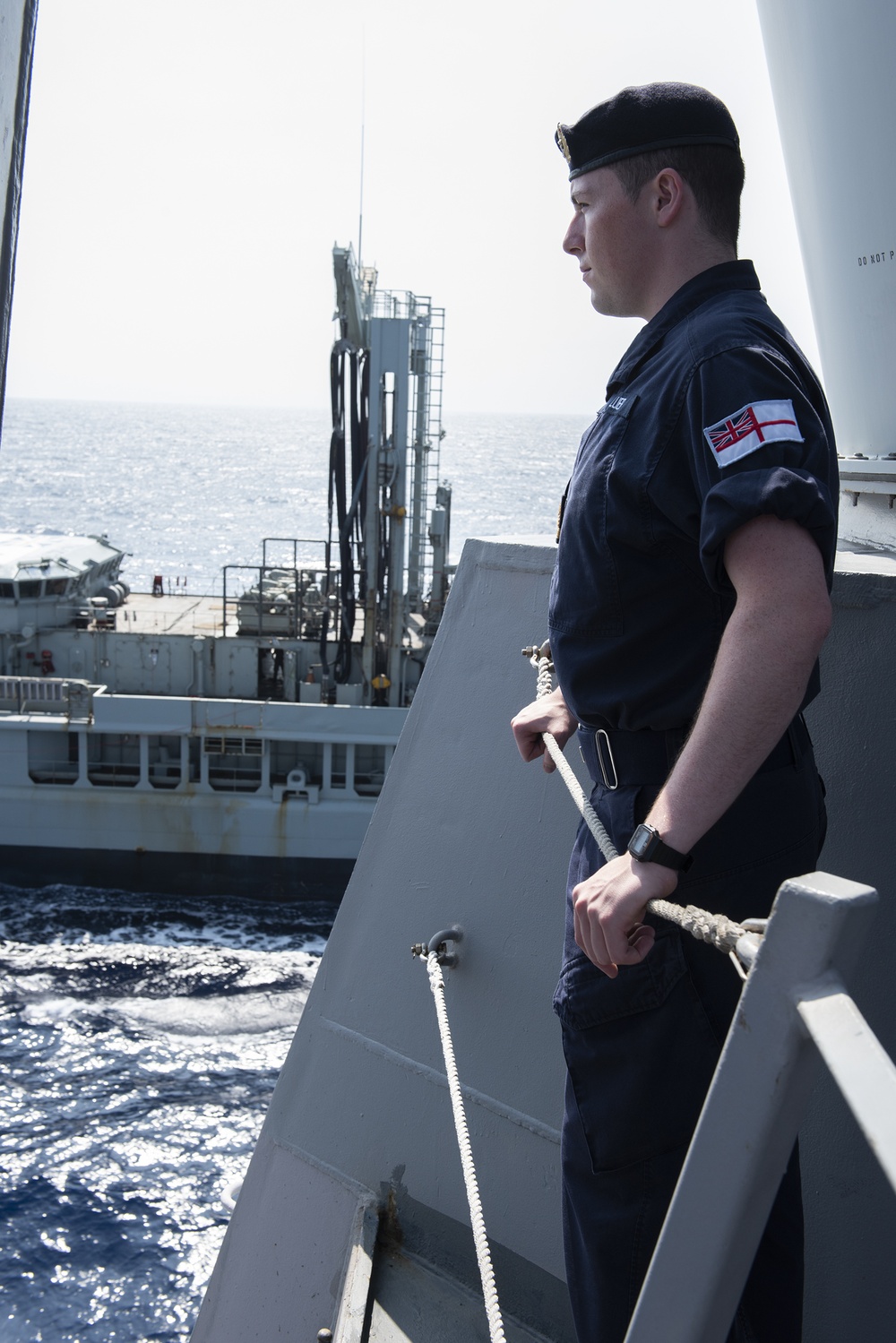 A Royal Navy sailor stands on the deck of a warship during what appears to be an underway replenishment (UNREP) operation with a support vessel alongside. The sailor is in profile view, wearing standard dark navy working dress uniform with a Royal Navy arm patch combining the Union Jack and White Ensign. He holds a rope/line and appears to be monitoring the replenishment operation.