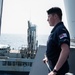 A Royal Navy sailor stands on the deck of a warship during what appears to be an underway replenishment (UNREP) operation with a support vessel alongside. The sailor is in profile view, wearing standard dark navy working dress uniform with a Royal Navy arm patch combining the Union Jack and White Ensign. He holds a rope/line and appears to be monitoring the replenishment operation.
