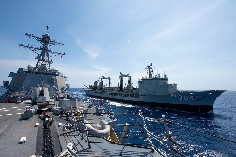 Two naval vessels are conducting an underway replenishment (UNREP) operation at sea, with transfer lines visibly connecting the two ships. The vessel on the right bears the hull number '304' with the prefix 'OR', indicating a replenishment or auxiliary ship. The ship on the left appears to be a guided-missile destroyer based on its superstructure, radar arrays, and deck-mounted weapons systems.