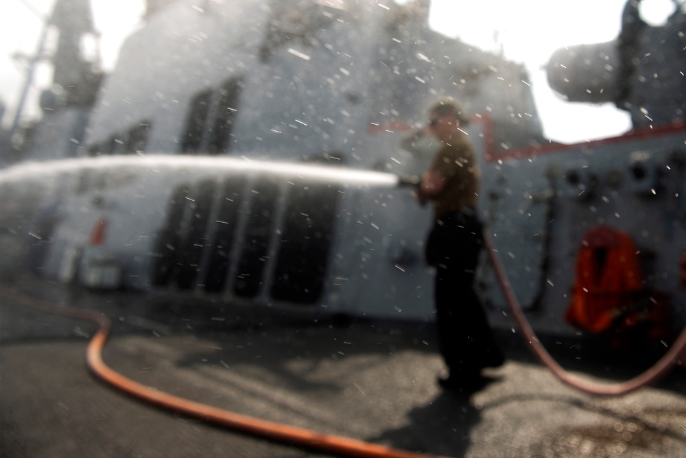 A person is hosing down the deck of a large naval or military vessel using an orange hose. Water spray is visible throughout the image, creating a mist effect. The scene appears to be routine deck maintenance or washdown operations aboard a warship.