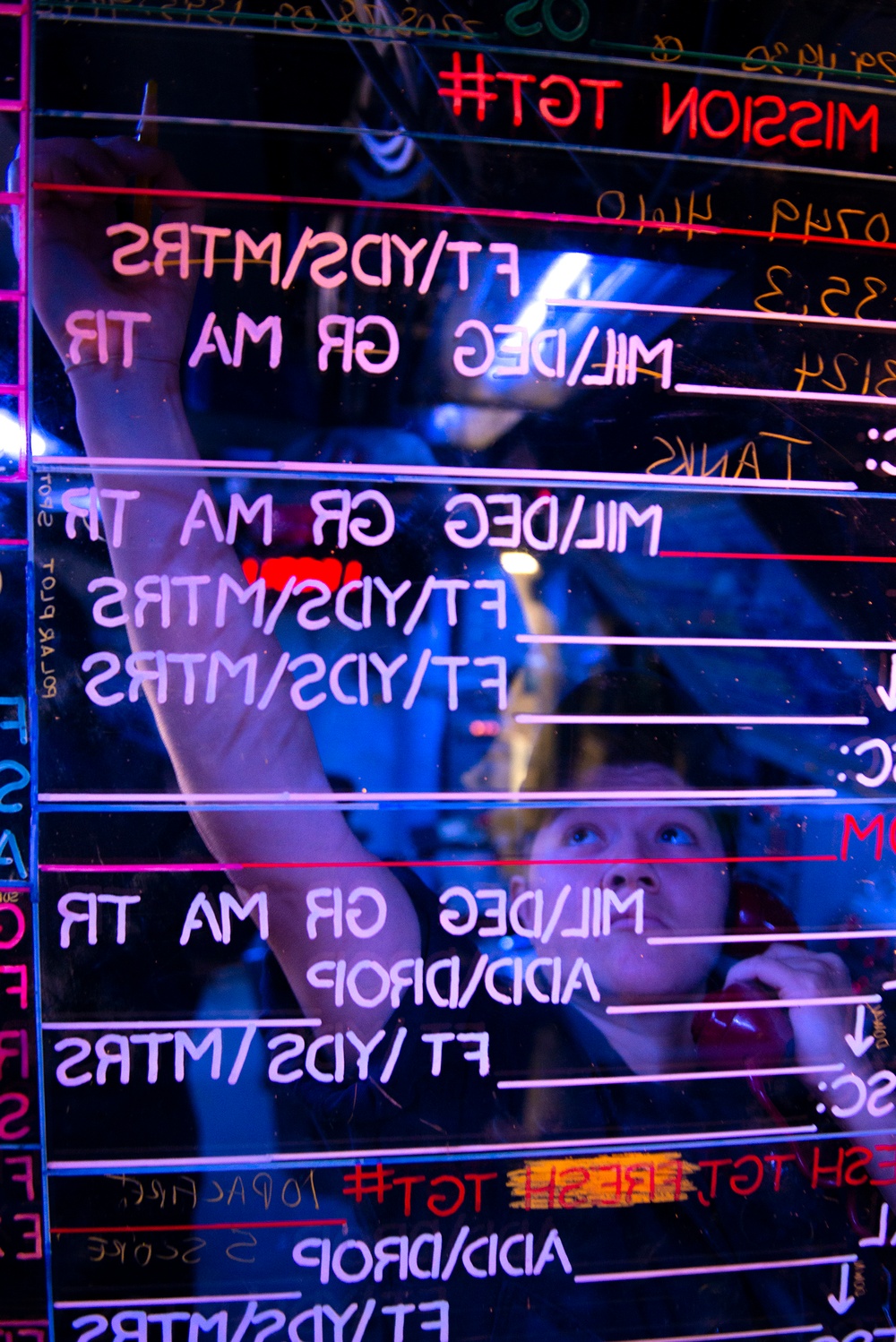 The image shows a person writing on a transparent glass or acrylic board with neon-lit text, photographed from the reverse side, causing all text to appear mirror-reversed. The board appears to be a military or tactical mission planning board with fields for measurements in FT/YDS/MTRS, MIL/DEG, GR, MA, TR, and ADD/DROP. The scene is illuminated with blue and pink neon lighting, creating a dramatic nightclub or gaming environment aesthetic.