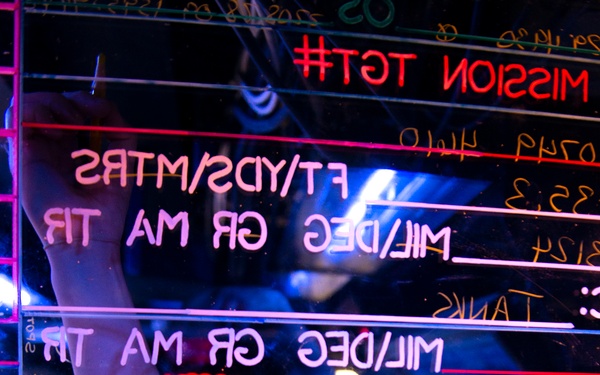 The image shows a person writing on a transparent glass or acrylic board with neon-lit text, photographed from the reverse side, causing all text to appear mirror-reversed. The board appears to be a military or tactical mission planning board with fields for measurements in FT/YDS/MTRS, MIL/DEG, GR, MA, TR, and ADD/DROP. The scene is illuminated with blue and pink neon lighting, creating a dramatic nightclub or gaming environment aesthetic.