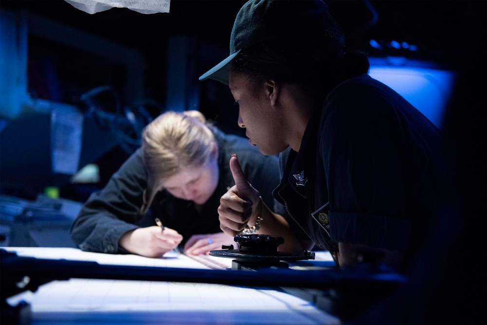 Two individuals are working together over an illuminated surface in a dimly lit environment, likely a naval vessel's chart room or bridge. One person is writing or marking on what appears to be a navigation chart, while the other, wearing a dark cap and uniform with an insignia patch, appears to be instructing or pointing. The blue-toned ambient lighting and navigation instruments suggest a military or maritime operational setting.