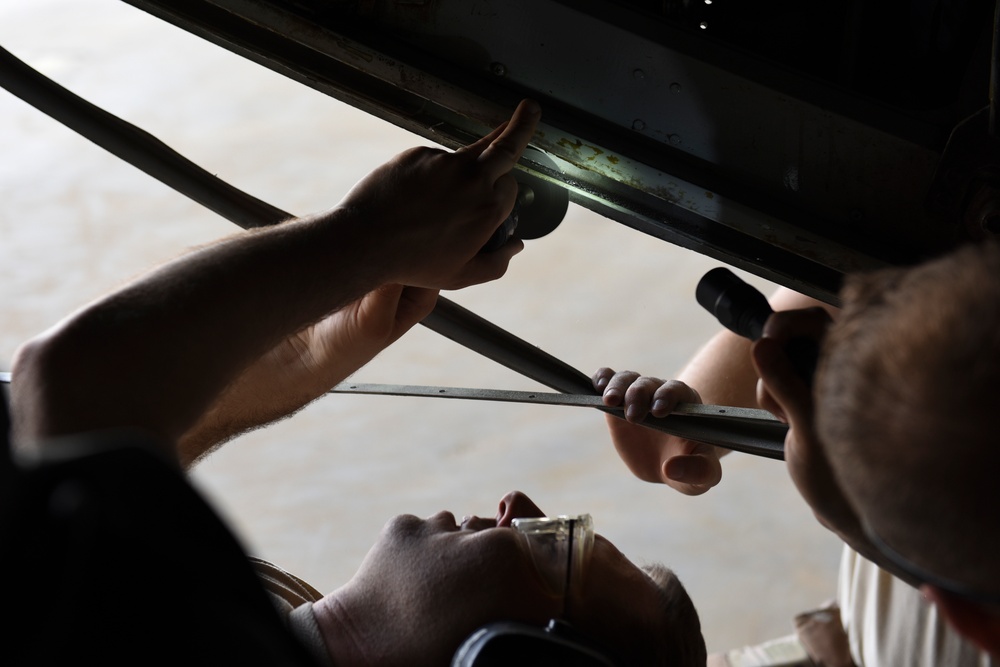 Two individuals are performing a mechanical inspection or repair task on what appears to be the underside of a vehicle or large equipment. One person is using a flashlight to illuminate a metal rail or track area while another uses a long-handled tool, possibly a wrench or ratchet extension. The scene appears to take place in a garage or workshop environment.