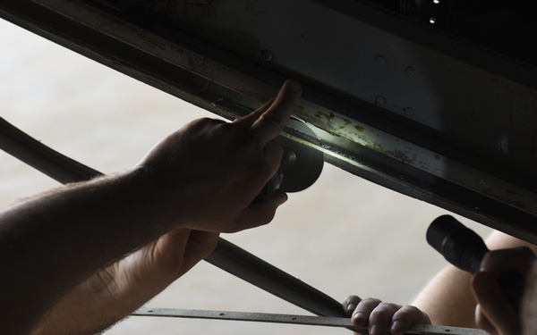 Two individuals are performing a mechanical inspection or repair task on what appears to be the underside of a vehicle or large equipment. One person is using a flashlight to illuminate a metal rail or track area while another uses a long-handled tool, possibly a wrench or ratchet extension. The scene appears to take place in a garage or workshop environment.