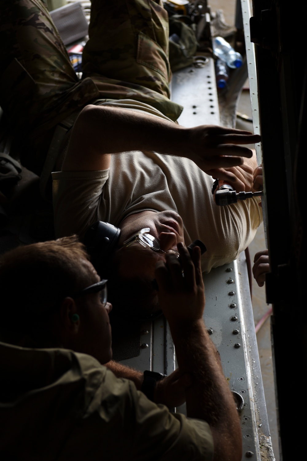 The image shows military personnel in camouflage uniforms performing maintenance or repair work on what appears to be a large military aircraft, likely working on an interior panel or structural component. The photo is rotated 90 degrees, making the subjects appear sideways. At least two individuals are visible using hand tools, with one wearing safety glasses.