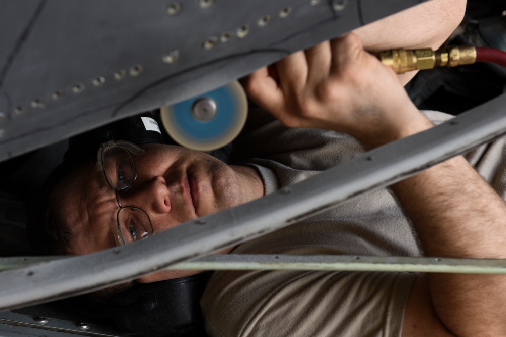 A technician or mechanic is working in a confined space beneath what appears to be an aircraft fuselage or wing structure. The individual is lying on their back, using a pneumatic grinding or polishing tool with a blue abrasive disc attached to an air hose with a brass fitting. The work environment shows riveted metal panels consistent with aircraft maintenance.