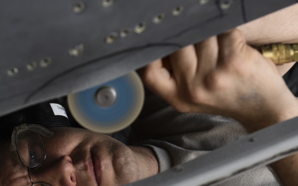 A technician or mechanic is working in a confined space beneath what appears to be an aircraft fuselage or wing structure. The individual is lying on their back, using a pneumatic grinding or polishing tool with a blue abrasive disc attached to an air hose with a brass fitting. The work environment shows riveted metal panels consistent with aircraft maintenance.