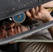 A technician or mechanic is working in a confined space beneath what appears to be an aircraft fuselage or wing structure. The individual is lying on their back, using a pneumatic grinding or polishing tool with a blue abrasive disc attached to an air hose with a brass fitting. The work environment shows riveted metal panels consistent with aircraft maintenance.