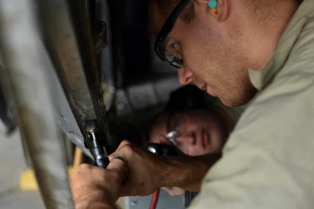 Two individuals are performing maintenance or repair work on what appears to be a metal structure or vehicle component. The foreground person is wearing safety glasses and ear protection while using a hand tool, likely a ratchet or socket wrench. A second person is visible in the background using a flashlight to illuminate the work area.