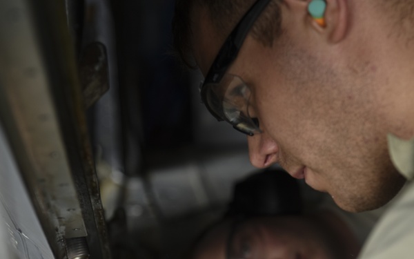 Two individuals are performing maintenance or repair work on what appears to be a metal structure or vehicle component. The foreground person is wearing safety glasses and ear protection while using a hand tool, likely a ratchet or socket wrench. A second person is visible in the background using a flashlight to illuminate the work area.