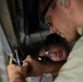 Two individuals are performing maintenance or repair work on what appears to be a metal structure or vehicle component. The foreground person is wearing safety glasses and ear protection while using a hand tool, likely a ratchet or socket wrench. A second person is visible in the background using a flashlight to illuminate the work area.