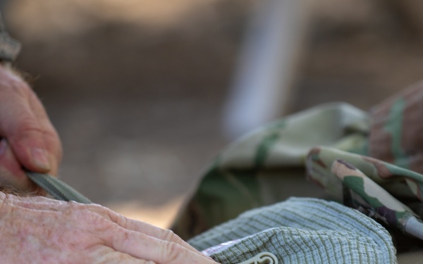 A close-up image showing hands in US Army ACU (Army Combat Uniform) digital camouflage applying what appears to be a field dressing or bandage to a casualty wearing OCP (Operational Camouflage Pattern) uniform. The focus is on the hands manipulating medical material near the torso area of the patient. This appears to be a tactical combat casualty care (TCCC) training or operational scenario.