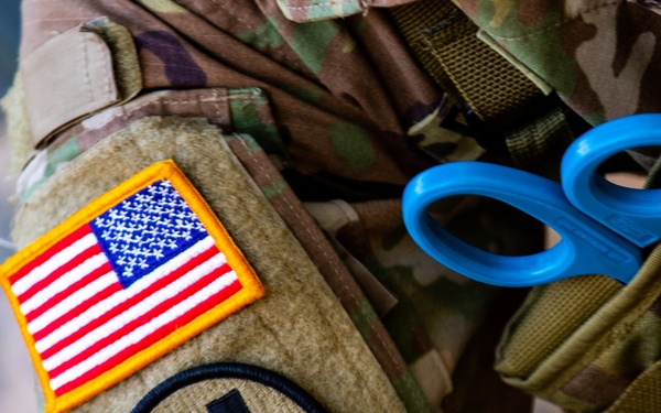 Close-up photograph of a U.S. military service member's upper arm and shoulder area in MultiCam/OCP camouflage uniform. The sleeve displays a full-color American flag patch with gold border and a circular unit insignia patch below it. Blue trauma scissors (shears) are visible clipped to gear on the right side of the frame.