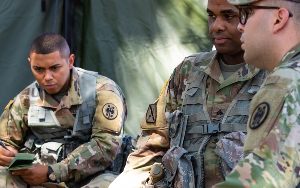 Three U.S. Army soldiers in Operational Camouflage Pattern (OCP) uniforms are gathered near a military tent in an outdoor setting. One soldier on the left is writing in a small notebook while the others appear to be engaged in discussion or observation. The scene suggests a field training exercise or operational briefing.