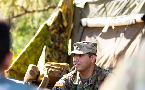 A U.S. Army soldier in MultiCam/OCP camouflage uniform is speaking to a group of personnel in an outdoor field setting near a military tent. The soldier appears to be conducting a briefing or instruction session, with blue scissors visible clipped to their uniform, suggesting a medical or combat lifesaver role. The background shows a tan military field tent with camouflage netting.