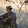 A male soldier in US Army Operational Camouflage Pattern (OCP) uniform is seated in dry grassland under sparse tree branches. He is wearing tactical sunglasses and a patrol cap, holding what appears to be a dark cylindrical object, and appears to be observing his surroundings. The setting suggests a field training exercise or operational pause in an arid environment.
