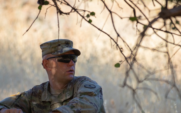 A male soldier in US Army Operational Camouflage Pattern (OCP) uniform is seated in dry grassland under sparse tree branches. He is wearing tactical sunglasses and a patrol cap, holding what appears to be a dark cylindrical object, and appears to be observing his surroundings. The setting suggests a field training exercise or operational pause in an arid environment.
